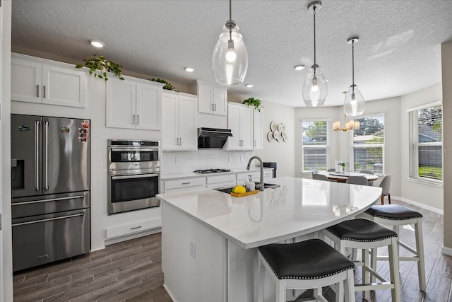 kitchen featuring pendant lighting, dark wood-type flooring, a center island with sink, appliances with stainless steel finishes, and white cabinetry