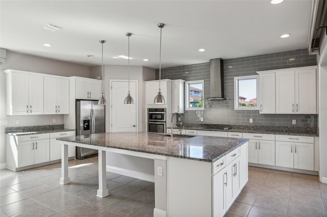 kitchen with white cabinetry, wall chimney exhaust hood, hanging light fixtures, a kitchen island with sink, and appliances with stainless steel finishes