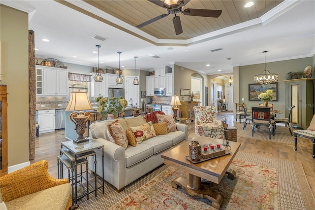 living room with ceiling fan with notable chandelier, light wood-type flooring, ornamental molding, and a tray ceiling