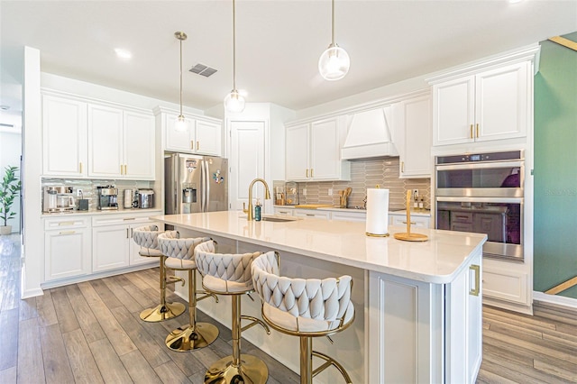 kitchen featuring light hardwood / wood-style flooring, custom range hood, appliances with stainless steel finishes, decorative light fixtures, and white cabinetry