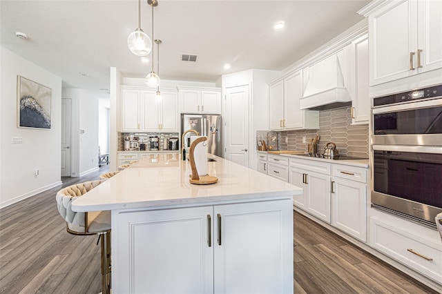 kitchen featuring appliances with stainless steel finishes, custom range hood, white cabinets, dark hardwood / wood-style floors, and an island with sink