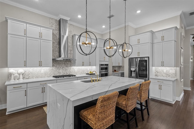 kitchen featuring wall chimney range hood, light stone counters, an island with sink, decorative light fixtures, and white cabinets