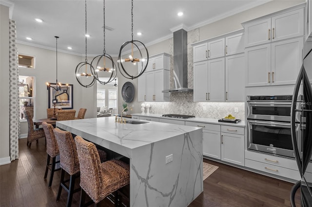 kitchen featuring white cabinetry, a center island with sink, decorative light fixtures, and wall chimney range hood