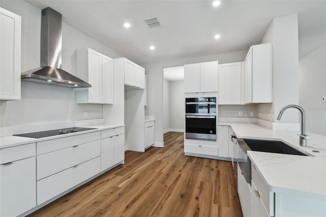 kitchen featuring wood finished floors, light stone countertops, multiple ovens, black electric cooktop, and wall chimney range hood