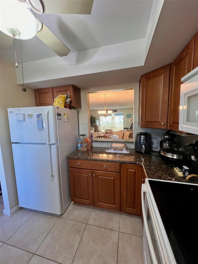 kitchen featuring ceiling fan with notable chandelier, light tile patterned flooring, dark stone counters, and white appliances