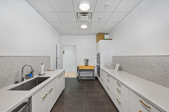 kitchen with a paneled ceiling, stainless steel dishwasher, sink, dark tile patterned flooring, and white cabinets