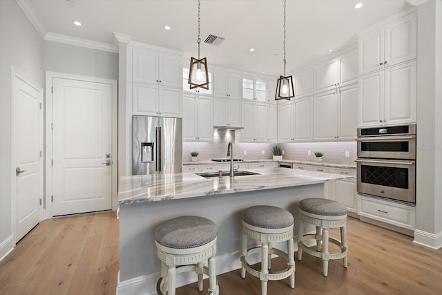 kitchen featuring light wood-type flooring, light stone counters, stainless steel appliances, white cabinets, and an island with sink