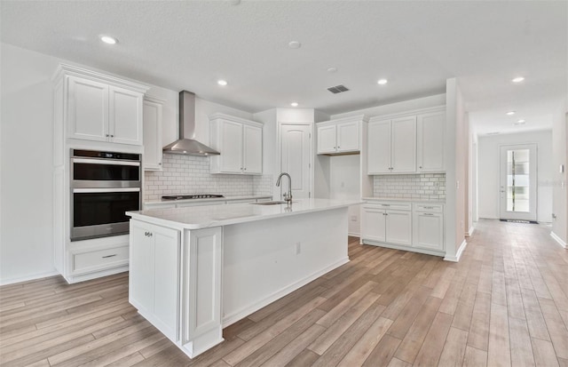 kitchen with white cabinets, a kitchen island with sink, sink, and wall chimney exhaust hood