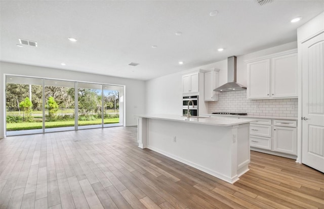 kitchen with white cabinets, light hardwood / wood-style flooring, a kitchen island with sink, and wall chimney range hood