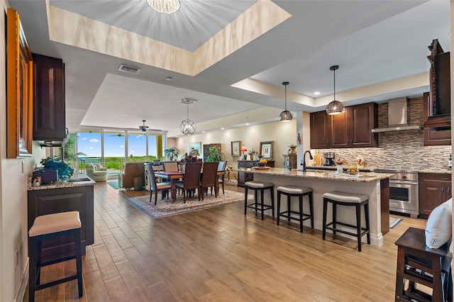 kitchen with wall chimney range hood, high end stainless steel range, light hardwood / wood-style flooring, an island with sink, and a kitchen bar