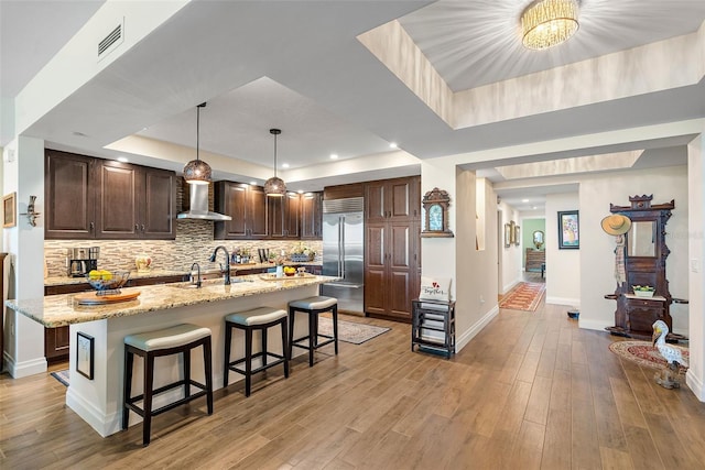 kitchen featuring light stone counters, a tray ceiling, light hardwood / wood-style flooring, built in refrigerator, and an island with sink