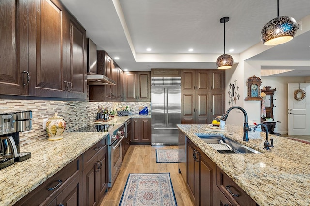 kitchen with light wood-type flooring, wall chimney exhaust hood, high end appliances, sink, and decorative light fixtures