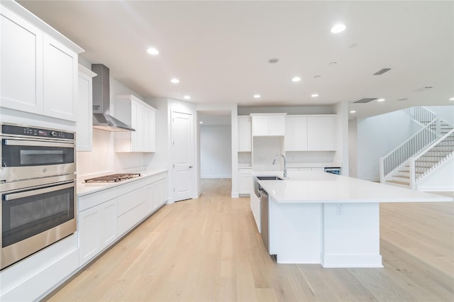 kitchen with a center island with sink, white cabinets, wall chimney range hood, light hardwood / wood-style flooring, and appliances with stainless steel finishes