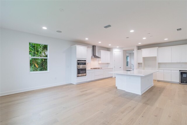 kitchen with wall chimney range hood, sink, light hardwood / wood-style flooring, an island with sink, and double oven