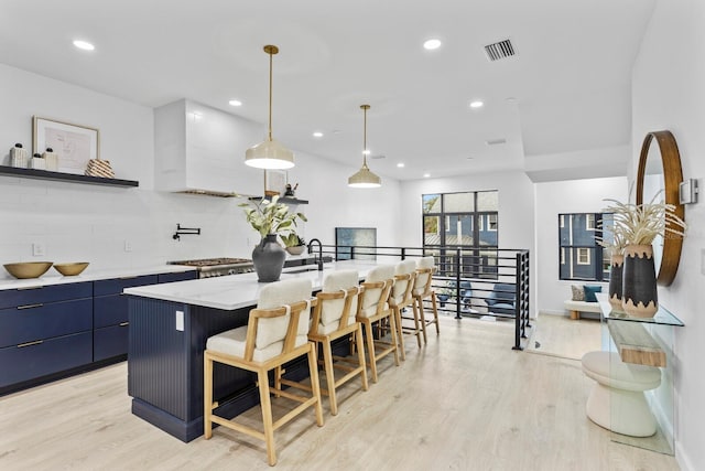 kitchen featuring tasteful backsplash, blue cabinets, decorative light fixtures, a kitchen island with sink, and light wood-type flooring