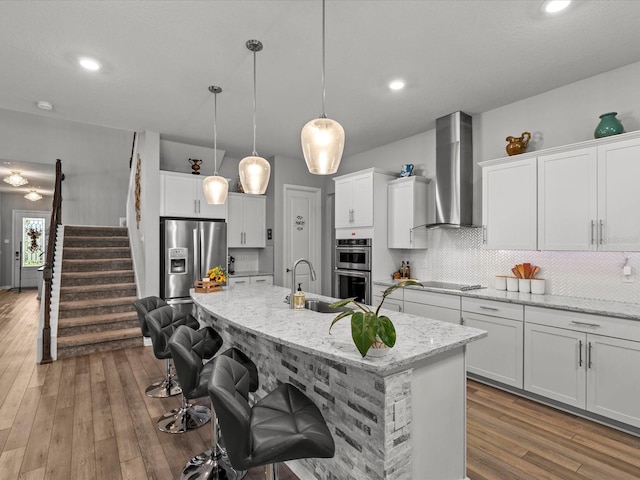 kitchen with appliances with stainless steel finishes, light wood-type flooring, wall chimney exhaust hood, sink, and pendant lighting