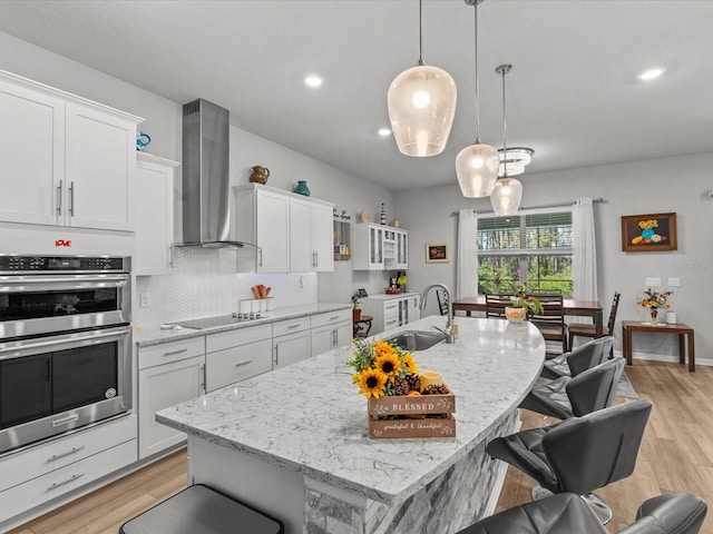 kitchen featuring wall chimney exhaust hood, white cabinetry, a kitchen island with sink, and double oven