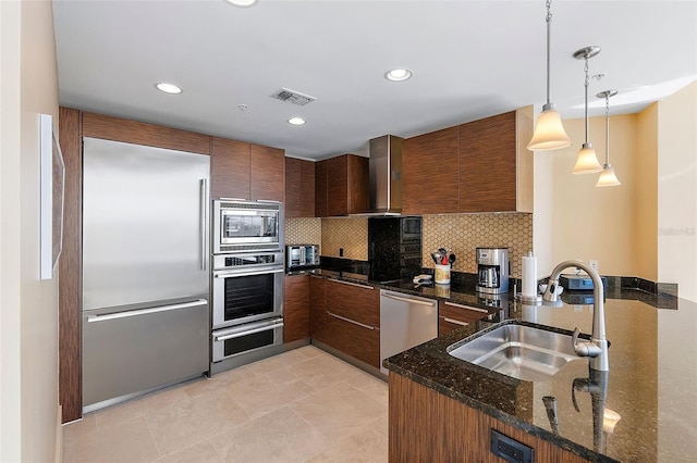 kitchen featuring dark stone counters, sink, wall chimney range hood, pendant lighting, and built in appliances