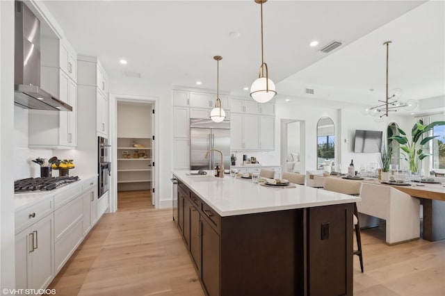 kitchen with stainless steel appliances, wall chimney range hood, pendant lighting, light hardwood / wood-style floors, and white cabinetry