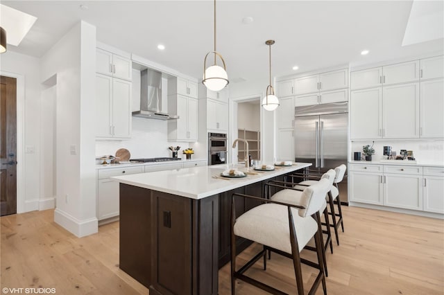 kitchen with light hardwood / wood-style floors, wall chimney exhaust hood, an island with sink, and appliances with stainless steel finishes