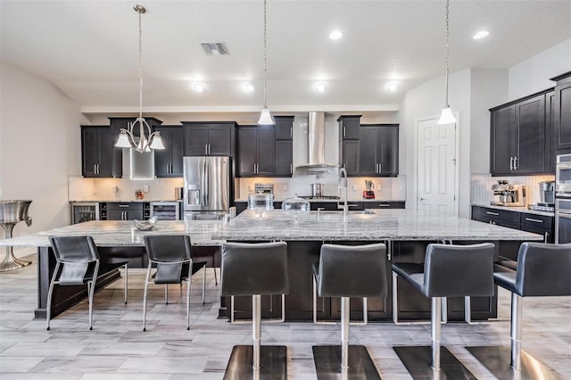 kitchen featuring wall chimney range hood, a large island with sink, decorative light fixtures, and stainless steel refrigerator with ice dispenser