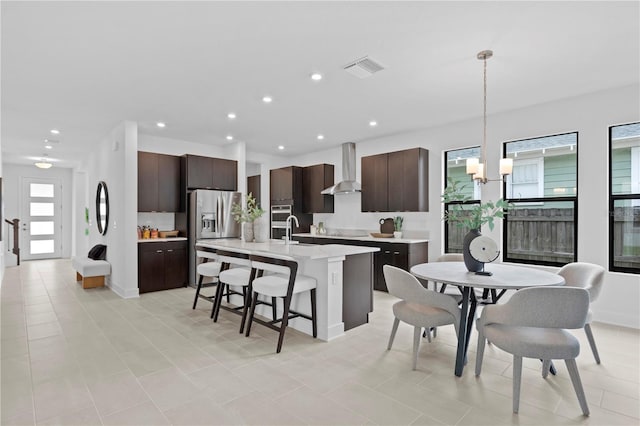 kitchen featuring wall chimney range hood, dark brown cabinetry, stainless steel appliances, and decorative light fixtures