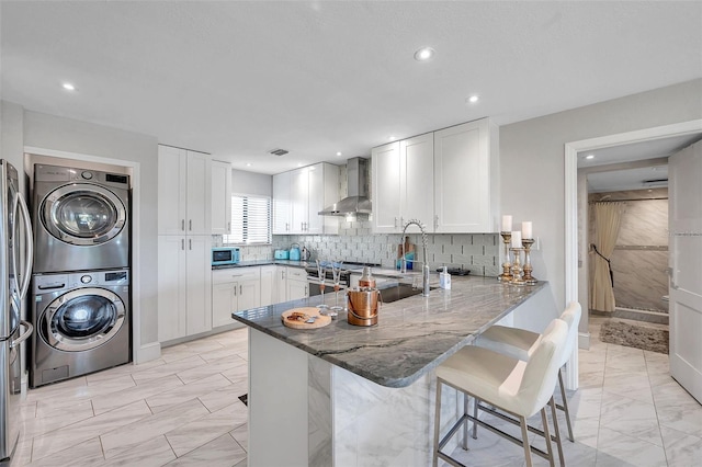 kitchen featuring stainless steel appliances, stacked washing maching and dryer, wall chimney range hood, a kitchen breakfast bar, and kitchen peninsula