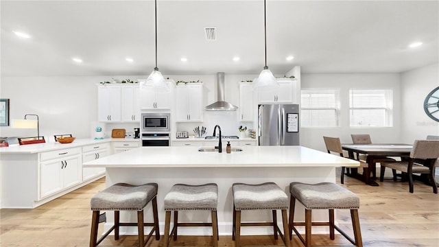 kitchen with hanging light fixtures, a center island with sink, wall chimney exhaust hood, and appliances with stainless steel finishes