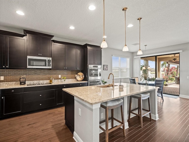 kitchen with stainless steel appliances, dark wood-type flooring, sink, decorative light fixtures, and an island with sink