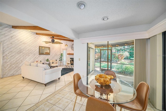 tiled dining space featuring wood walls, a stone fireplace, ceiling fan, a textured ceiling, and beam ceiling