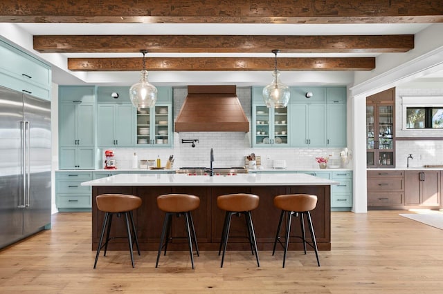 kitchen featuring backsplash, custom exhaust hood, beam ceiling, an island with sink, and built in fridge