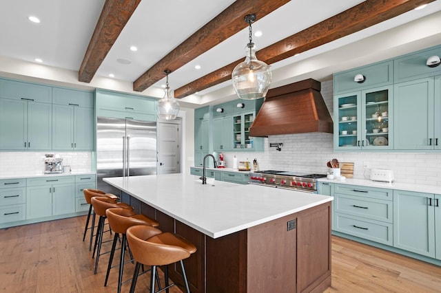 kitchen with custom exhaust hood, beamed ceiling, hanging light fixtures, a kitchen island with sink, and high quality appliances