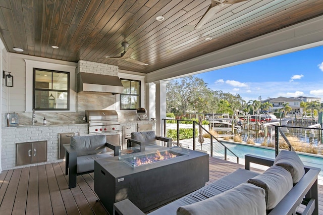 wooden terrace featuring an outdoor living space with a fire pit, ceiling fan, exterior kitchen, a grill, and a water view