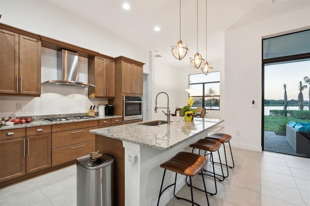 kitchen with a kitchen island with sink, sink, wall chimney exhaust hood, light stone countertops, and appliances with stainless steel finishes