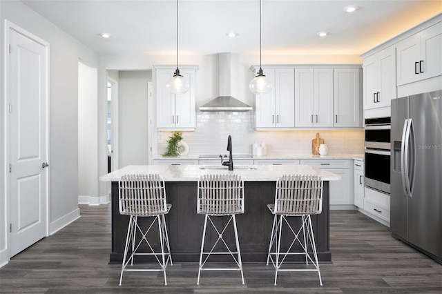 kitchen with appliances with stainless steel finishes, pendant lighting, a center island with sink, and wall chimney exhaust hood