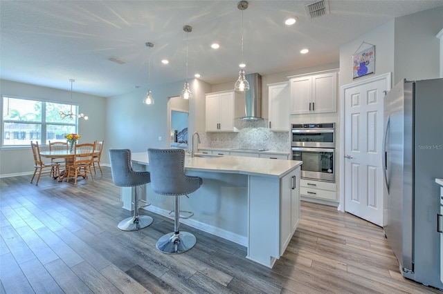 kitchen with sink, hanging light fixtures, stainless steel appliances, wall chimney range hood, and white cabinets