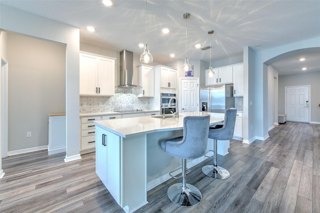 kitchen with a center island with sink, wall chimney exhaust hood, decorative light fixtures, white cabinetry, and wood-type flooring