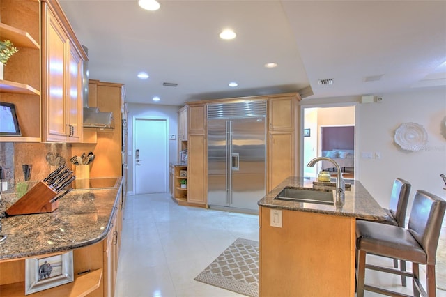 kitchen with a kitchen breakfast bar, backsplash, dark stone counters, stainless steel built in refrigerator, and sink