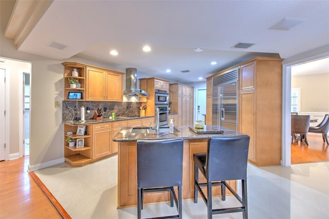 kitchen with a breakfast bar, a center island with sink, wall chimney exhaust hood, light wood-type flooring, and appliances with stainless steel finishes