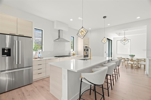 kitchen with a center island with sink, light hardwood / wood-style flooring, wall chimney exhaust hood, decorative light fixtures, and stainless steel appliances