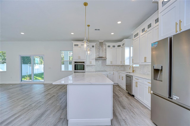 kitchen featuring wall chimney range hood, hanging light fixtures, appliances with stainless steel finishes, a kitchen island, and white cabinetry