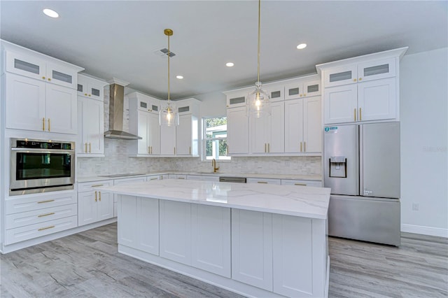 kitchen with appliances with stainless steel finishes, a kitchen island, wall chimney range hood, white cabinetry, and hanging light fixtures