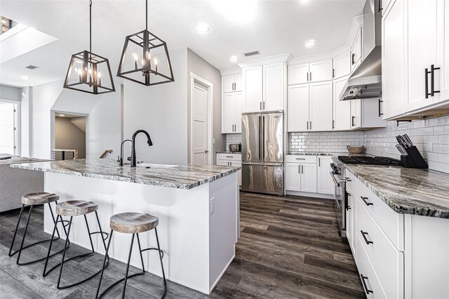 kitchen with wall chimney exhaust hood, dark wood-type flooring, hanging light fixtures, a kitchen island with sink, and high quality appliances