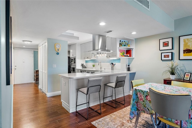 kitchen with wall chimney exhaust hood, dark wood-type flooring, kitchen peninsula, a breakfast bar, and white cabinets