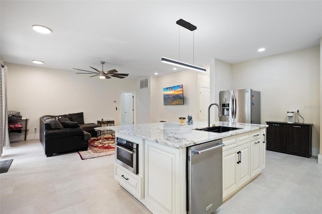 kitchen with light stone counters, a ceiling fan, a center island with sink, stainless steel appliances, and a sink