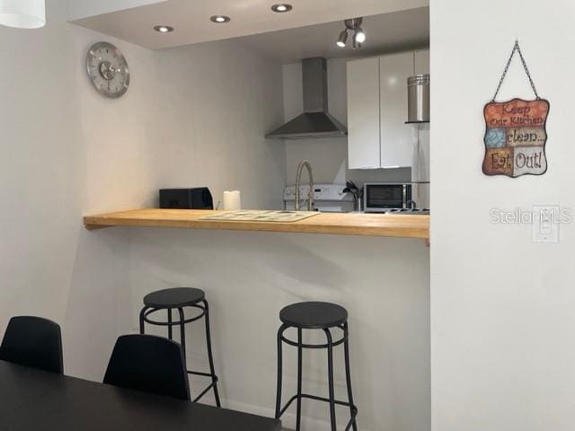 kitchen featuring a breakfast bar, kitchen peninsula, sink, wall chimney exhaust hood, and white cabinetry