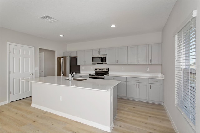 kitchen with sink, gray cabinets, a kitchen island with sink, appliances with stainless steel finishes, and light wood-type flooring