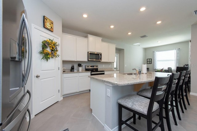 kitchen with light stone counters, stainless steel appliances, sink, white cabinets, and an island with sink