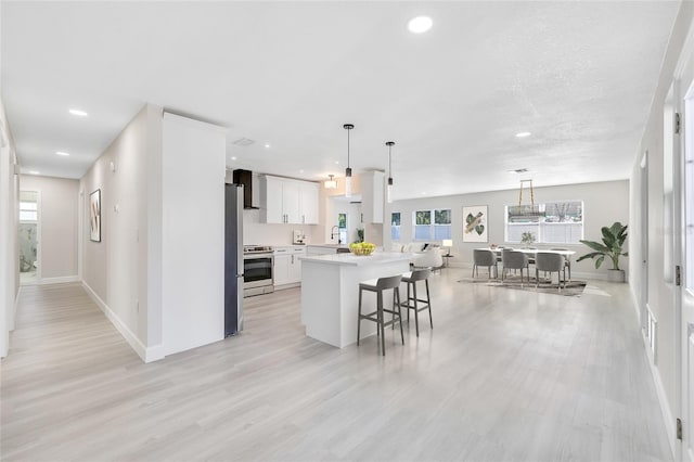 kitchen with light wood-type flooring, wall chimney exhaust hood, stainless steel appliances, pendant lighting, and white cabinets