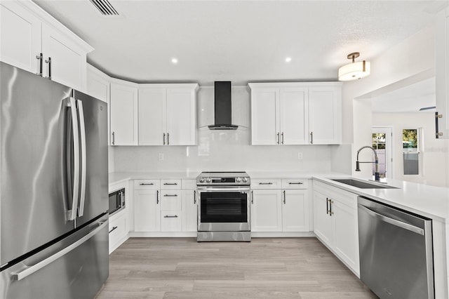 kitchen with appliances with stainless steel finishes, wall chimney exhaust hood, sink, white cabinets, and light hardwood / wood-style floors